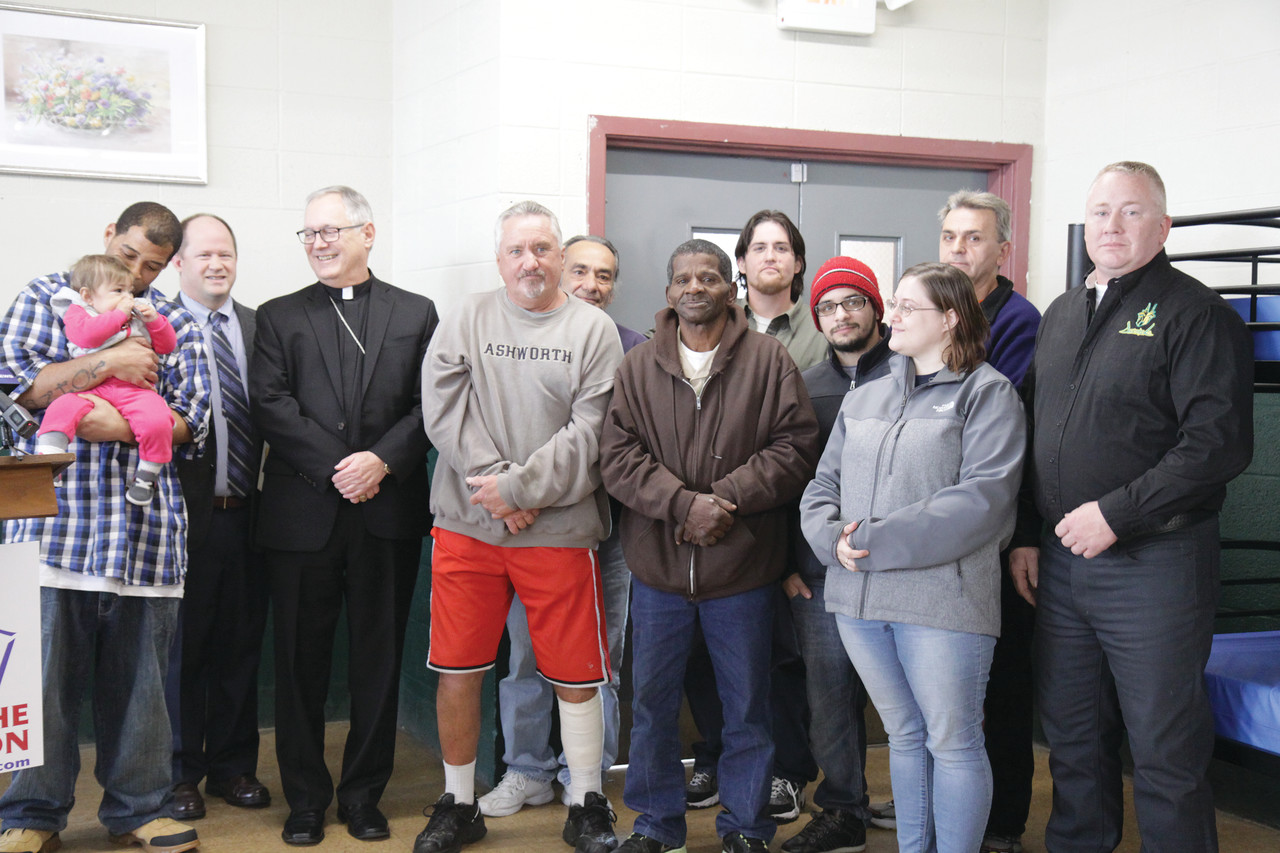 STANDING TOGETHER FOR THOSE IN NEED: Residents and former residents of Emmanuel House homeless shelter stand with Bishop Thomas J. Tobin and diocesan Emergency Services Coordinator James Jahnz (second from left) after making a donation to the “Keep the Heat On” campaign at Monday’s launch ceremony. Joe Patenaude (far right), a representative of Anubis LTD, a company that hires many residents of the shelter through a work partnership program, also presented a donation to the campaign.