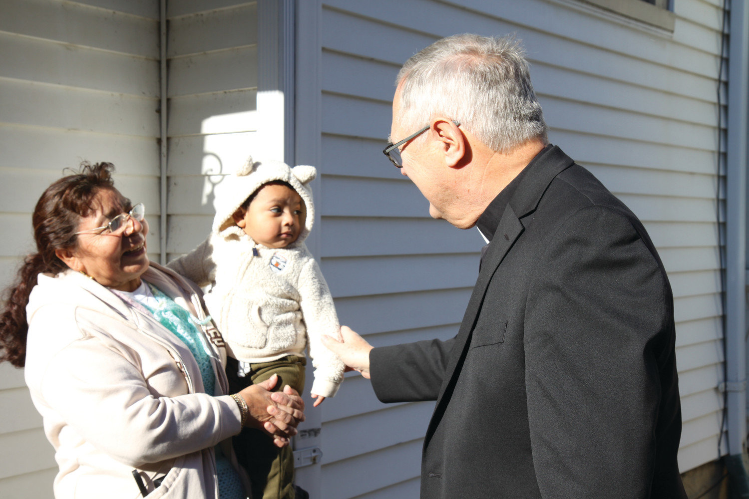 In this 2017 photo, Bishop Thomas J. Tobin greets one-year-old Liam Andres and his grandmother, Consuelo Robles, outside the home of Liam’s parents, Otoniel Andres and Natalia Zapata. The family was a beneficiary of the “Keep the Heat On” diocesan emergency heating assistance program.